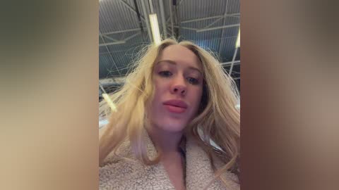 A video of a young, fair-skinned woman with long, blonde hair, wearing a fluffy beige bathrobe, taken indoors with a metal door in the background.