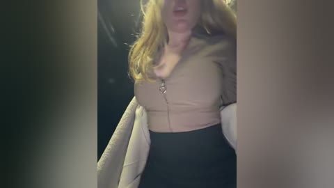 Video of a fair-skinned woman with long, blonde hair, wearing a beige, zip-up top and black skirt, partially obscured by dark, blurry background.