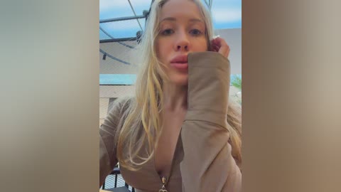 A video of a blonde woman with long hair, wearing a beige jacket, holding a brown object to her mouth, in an outdoor setting with a building and blue sky in the background.