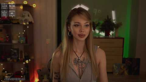 Video of a slender, fair-skinned woman with long blonde hair, wearing a low-cut, grey top with a necklace, standing in a dimly lit room with green lights, shelves, and a dresser.