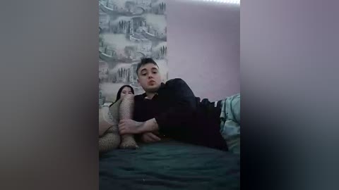 A video of a young Asian man lying on a bed, wearing a black hoodie, with a cat resting on his chest. The room has gray wallpaper and pastel-colored bedding.