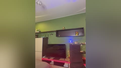 Video of a modern living room with a green wall, black floating shelves, a large flat-screen TV, and a red LED strip under a white desk.