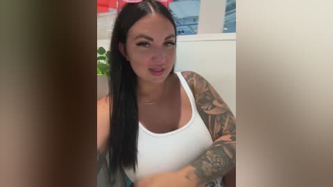 Video of a fair-skinned woman with long black hair, wearing a white tank top and pink headband, displaying tattoos on her arms. She stands indoors with blurred, white background.