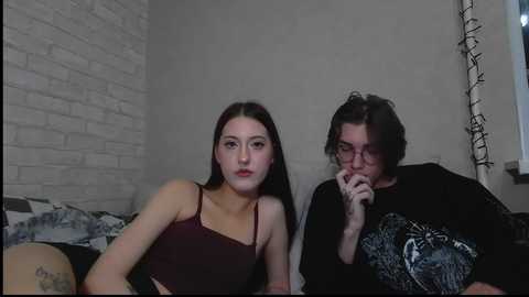 Video of a young woman with pale skin and long dark hair, wearing a burgundy crop top, sitting on a bed in a dimly lit room with a brick wall background and string lights.