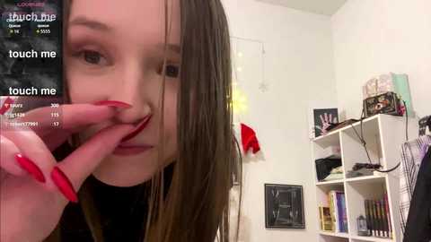 A young woman with straight, long brown hair and red nails touches her nose, wearing a black top. The background features a white wall with a bookshelf and a red coat hanging. The image has a video overlay.