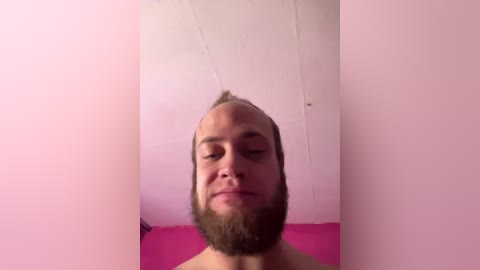 Video of a bald, bearded man with a spiky hair tuft, wearing nothing, standing against a pink wall with visible ceiling joints.