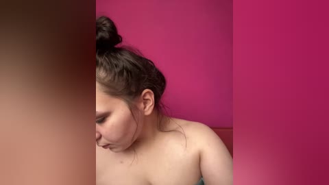 Video of a young woman with light skin, dark hair in a messy bun, wearing a strapless top, against a vibrant pink background, cropped to focus on her face and upper body.