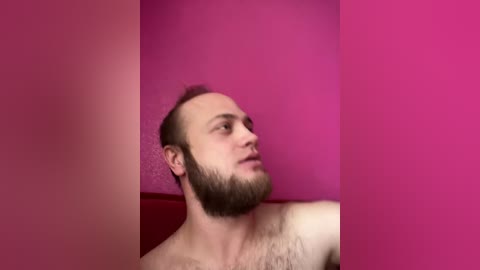 Video of a Caucasian man with a short beard, light skin, and a shaved head, wearing a red shirt, against a pink background. He appears contemplative.