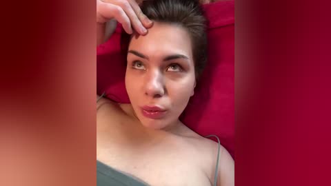 Video of a young woman with light skin and brown hair lying on a red cushioned surface, wearing a green tank top, with a hand touching her forehead, creating a serene, intimate atmosphere.