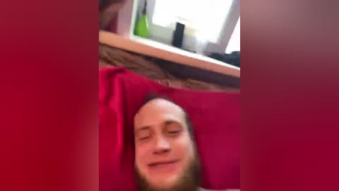 A blurry, candid video of a smiling, bald man lying on a red pillow, with a window in the background, creating a warm, cozy indoor setting.