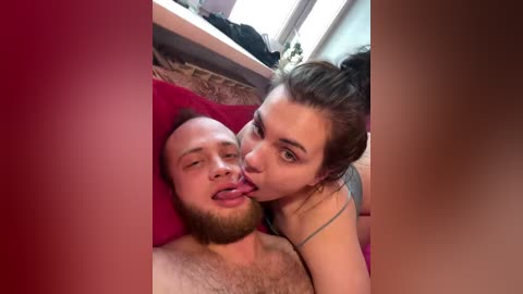 A candid video of a topless man with a beard and a woman with brown hair licking his face, both wearing casual clothes, in a dimly lit room with red cushions and a window.