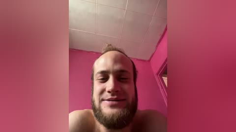 Video of a smiling, balding man with a beard, standing shirtless against a pink wall with a white ceiling.