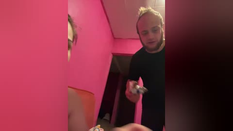 Video of a young woman with blonde hair, wearing a black top, holding a gun, standing in a pink room with a wooden door in the background.