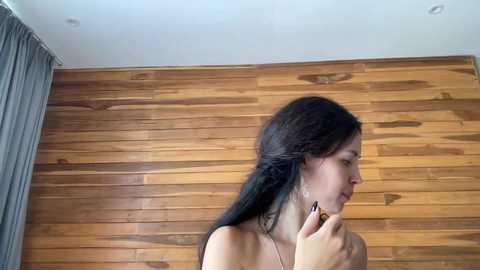 Video of a young woman with long dark hair, light skin, and a slender build, facing left, standing against a wooden paneled wall, with a gray curtain on the left.