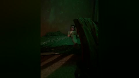 A video captures a man in a green t-shirt and jeans sitting on a wooden crate, partially obscured by a dark, shadowy figure in the foreground. The setting is dimly lit, with a murky green background, creating an eerie, suspenseful atmosphere.