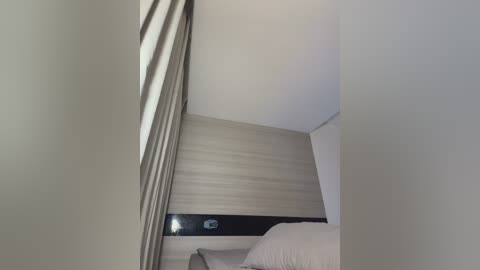 A video of a minimalist bedroom with a beige wooden headboard, white bedding, and a vertical black accent strip. The image is taken from a low angle, showing the ceiling and wall textures.