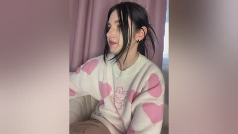 Video of a woman with long black hair, wearing a white sweater with pink hearts, sitting on a beige couch with pink curtains behind her.