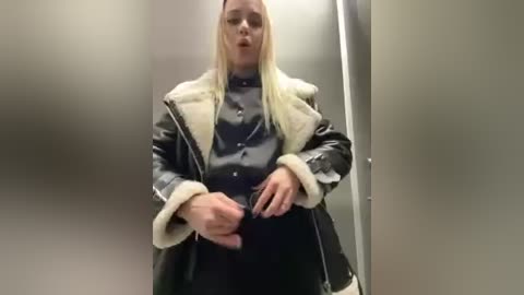 Video of a blonde woman in a black leather jacket with fur trim, standing in a dimly lit, beige-walled hallway, holding a smartphone.