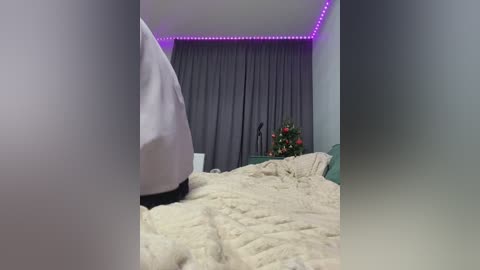 Video of a dimly lit bedroom with a gray curtain backdrop, a cozy white blanket, and a small Christmas tree with red and green decorations on the right.
