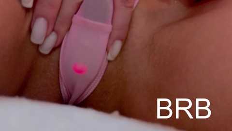Close-up video of a woman's shaved pubic region with light skin tone. She is wearing sheer, pink lingerie with a small heart emblem. Her neatly manicured hand is gently spreading her labia. \"BBR\" logo in white text is visible in the lower right corner.