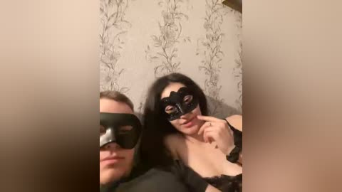 Video of two women in black masquerade masks, one with long black hair and a black bra, the other with blonde hair and a black bra, against a floral wallpaper background.