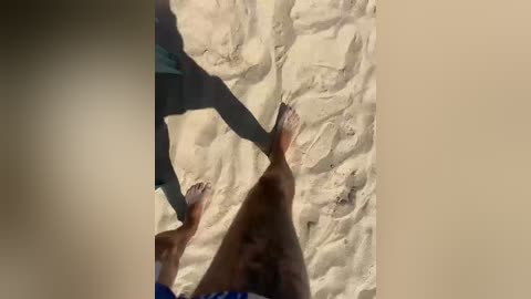 A video of a sandy beach with a woman's legs in blue shorts and a man's legs in shorts and a shirt, casting shadows on the sand.
