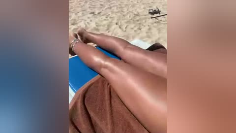 Video of a woman's legs, tan and smooth, lying on a towel with a blue edge, on a sandy beach with a clear blue sky background.