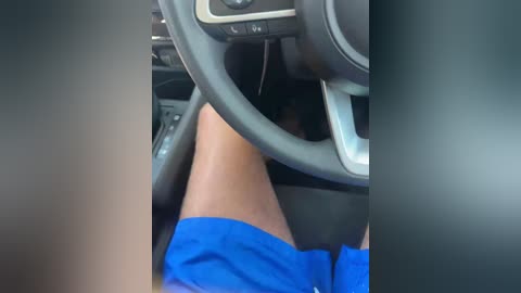 Video of a person's legs in blue shorts, sitting in a car with a gray dashboard and steering wheel.