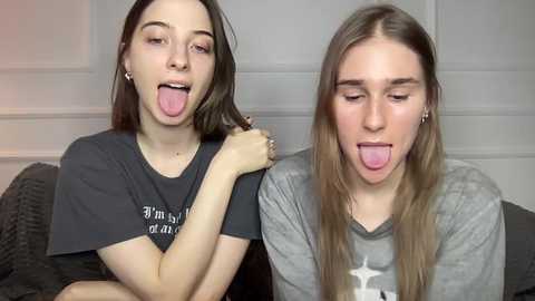 Video of two young women with long hair, wearing casual gray t-shirts, sticking out their tongues playfully. Background shows a gray couch and white wall.