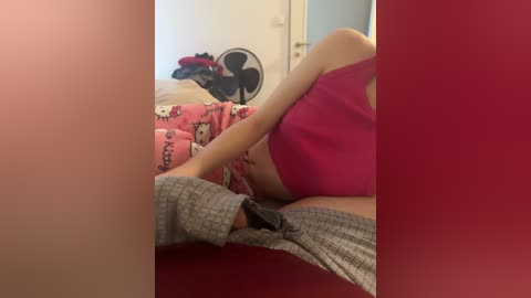 Video of a woman in a red tank top and gray pants lying on a bed with a pink Hello Kitty duvet.