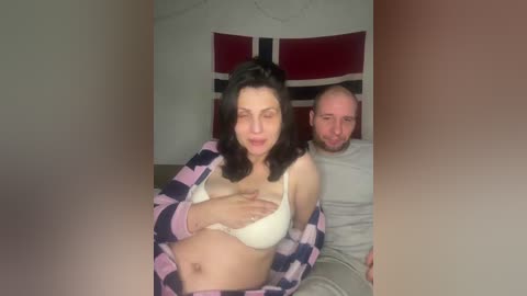 Video of a pregnant woman with light skin, dark hair, and a white bra, lying on a bed, smiling. Behind her, a bald man with a beard and grey shirt sits.