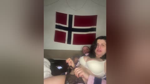 A video of a middle-aged man and woman in a dimly lit room. The woman, with medium breasts, is partially undressed, holding a bottle. The man, wearing a suit, sits behind her. A Norwegian flag hangs on the wall.