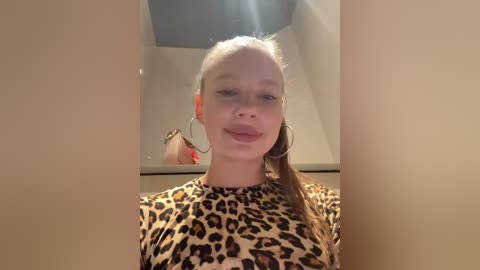 Video of a young, fair-skinned woman with straight, light brown hair, wearing large hoop earrings, and a leopard-print shirt, smiling softly. Background features a beige wall and a small mirror.