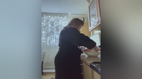 A video of a woman in a black dress standing at a kitchen counter, preparing food, with a white curtain and wooden cabinets in the background.