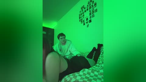 A video of a man lying on a bed with a patterned quilt, wearing a hoodie and shorts, under greenish lighting. The room features a collage of black-and-white photos on a wall.