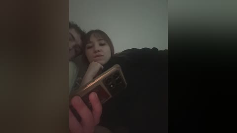 Video of a young woman with short brown hair, eyes closed, leaning on a man's shoulder, holding a phone. Background is dark and blurry.