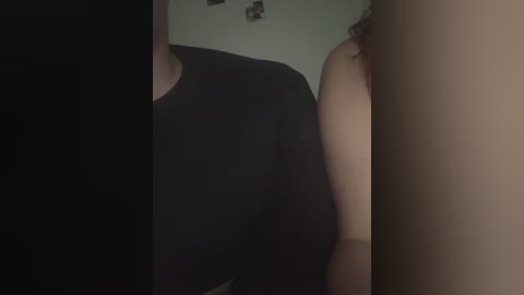 Video of a dimly lit room showing a person in a black shirt, partially obscured by a beige wall with a small, indistinct image. The background is out of focus.