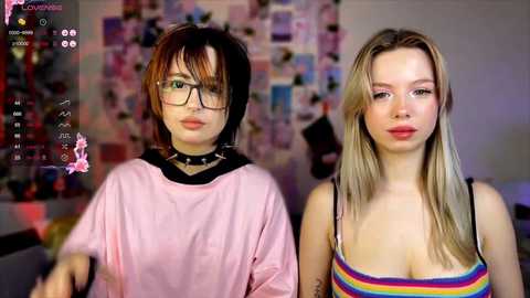 Video of two young women with pale skin, one in glasses and a black choker, the other with long blonde hair, rainbow-striped tank top. Background features a cluttered wall with photos.
