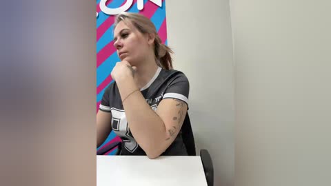 Video of a young blonde woman in a grey t-shirt, leaning on a table with her chin in her hand, against a colorful striped wall in a minimalist room.