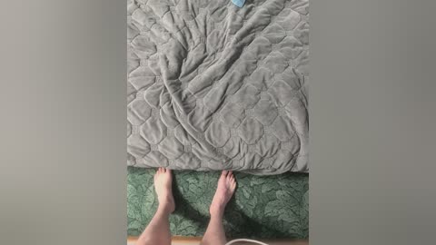 A video from an overhead perspective shows two bare feet on a green quilted bedspread, above a gray quilted blanket. The feet are light-skinned, and the image has a neutral color palette.