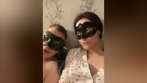 Video of two women in black Venetian masks, one with dark hair and white floral dress, the other with blonde hair, both indoors with textured wall background.