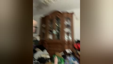 A blurred video of a living room with a wooden cabinet, a colorful blanket, and indistinct furniture, possibly a TV or another piece of furniture, in the background. The room has neutral beige walls and appears to be dimly lit.