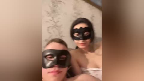 Video of two young women, one with light skin and blonde hair, the other with darker skin and dark hair, both wearing black masquerade masks, seated on a beige couch with floral-patterned cushions.