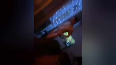 Video of a dimly lit room with a blurred man in underwear standing near a glowing, abstract blue light display on a wall. The image has a grainy texture and a soft focus, suggesting a nighttime setting.