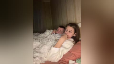 A video captures a couple in bed, with a woman in a white robe and a man in white pajamas, both sleeping, under a white fur blanket, in a dimly lit room.