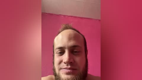 Video of a balding man with a receding hairline, a light beard, and a closed-mouth smile against a bright pink wall.