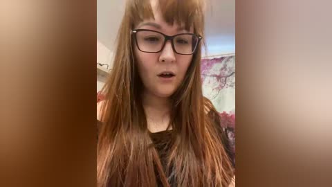 A video of a young woman with long, straight brown hair, wearing black-rimmed glasses, standing in front of a blurred, colorful background. She has a neutral expression.