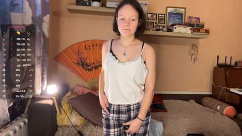Video of a young woman with short black hair, wearing a white camisole and plaid pajama pants, standing in a cluttered, dimly-lit bedroom with shelves of framed photos, a fan, and a TV screen displaying a video feed.