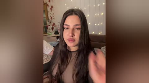 Video of a young woman with long black hair, wearing a black mesh top, puckering her lips, in a cozy bedroom with floral wallpaper, a bed, and string lights in the background.