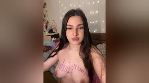 Video of a topless young woman with long dark hair, fair skin, and small breasts, with \"the only reason\" written on her chest. Background features a cozy bedroom with a bed, string lights, and floral decorations.